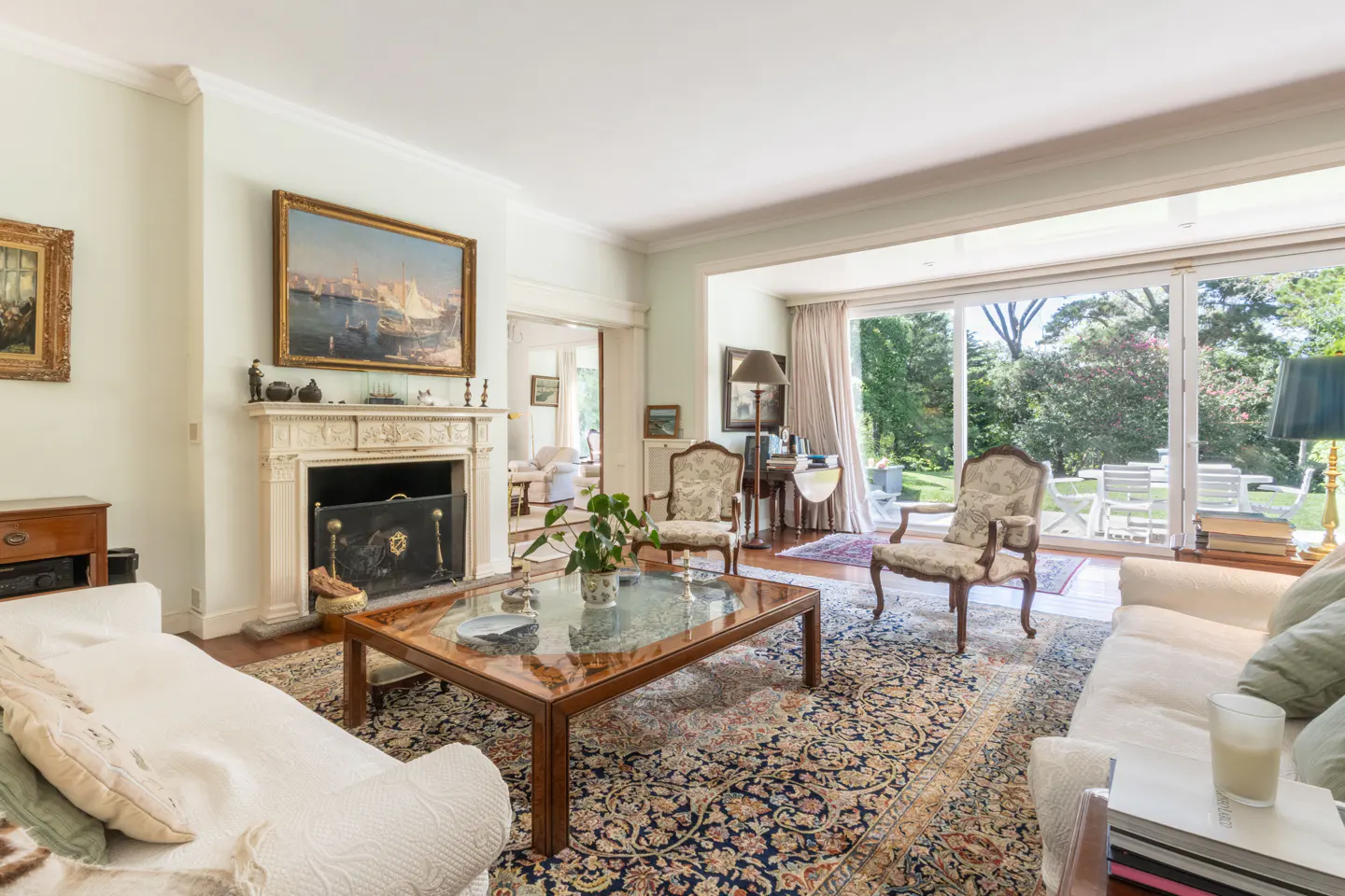 Bright living room with white sofas, floral chairs, and a large rug. A fireplace and sliding glass doors to a green yard add charm.