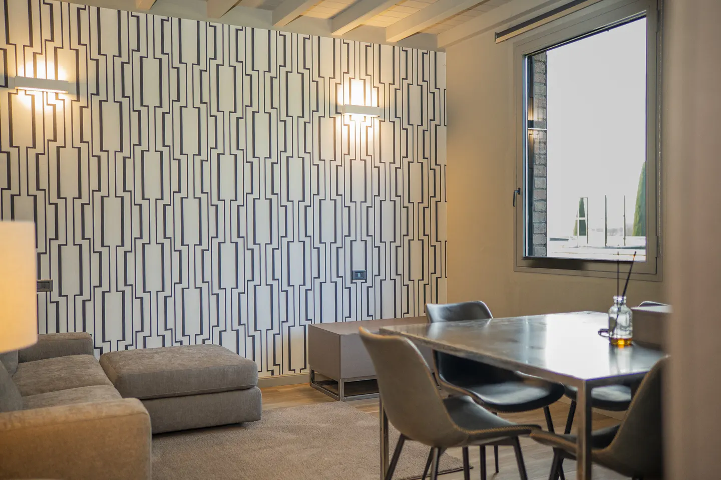 Living room with a gray sofa, ottoman, and a dining table with four chairs. The wall has a black and white geometric pattern. A window overlooks a rooftop.
