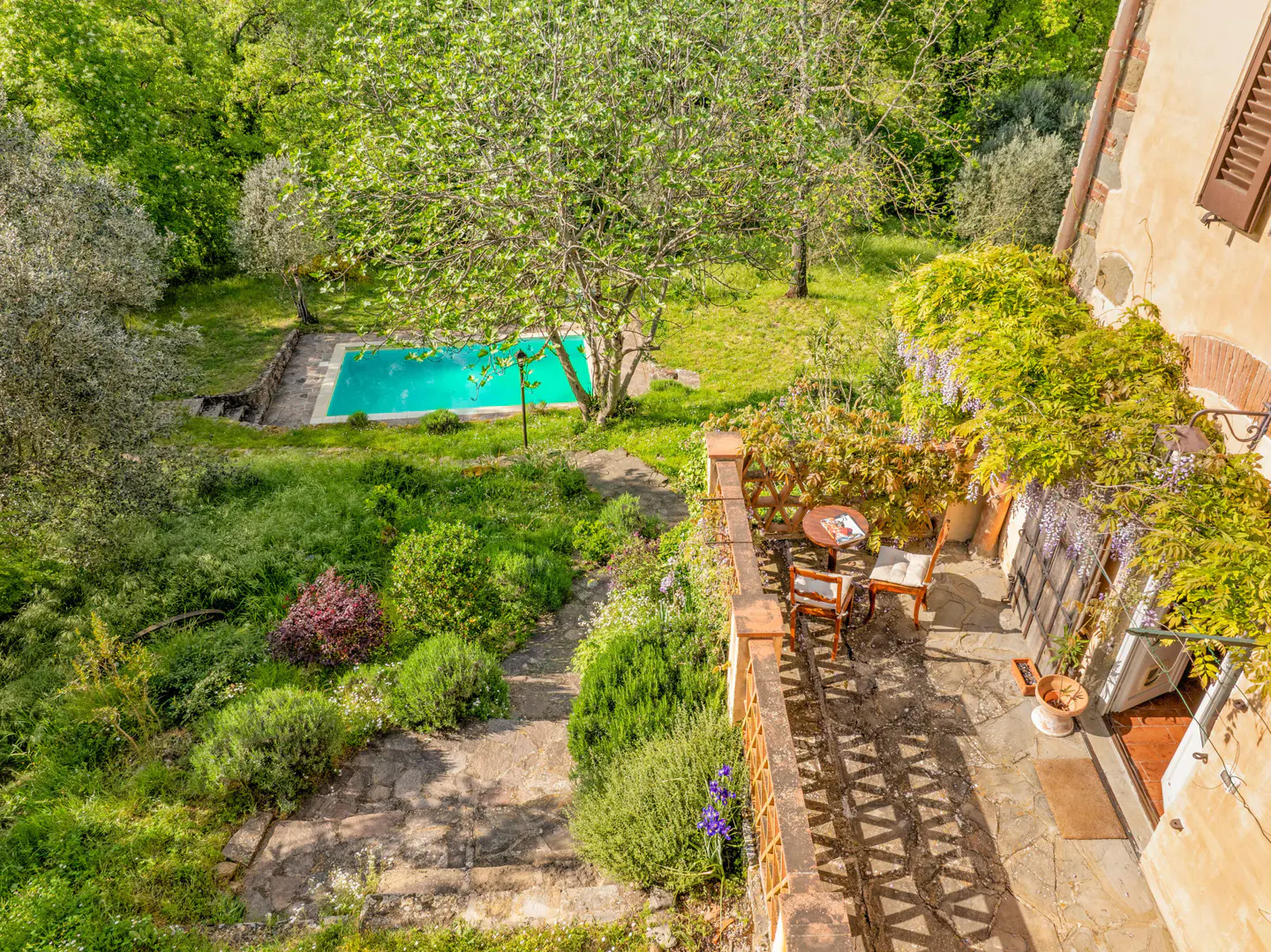 Overhead view of a stone patio with table and chairs, wisteria, and a pool in the background.