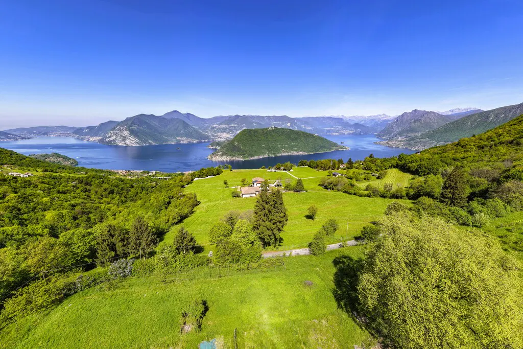 Aerial view of a green hillside with a house, overlooking a blue lake and mountains under a clear blue sky.