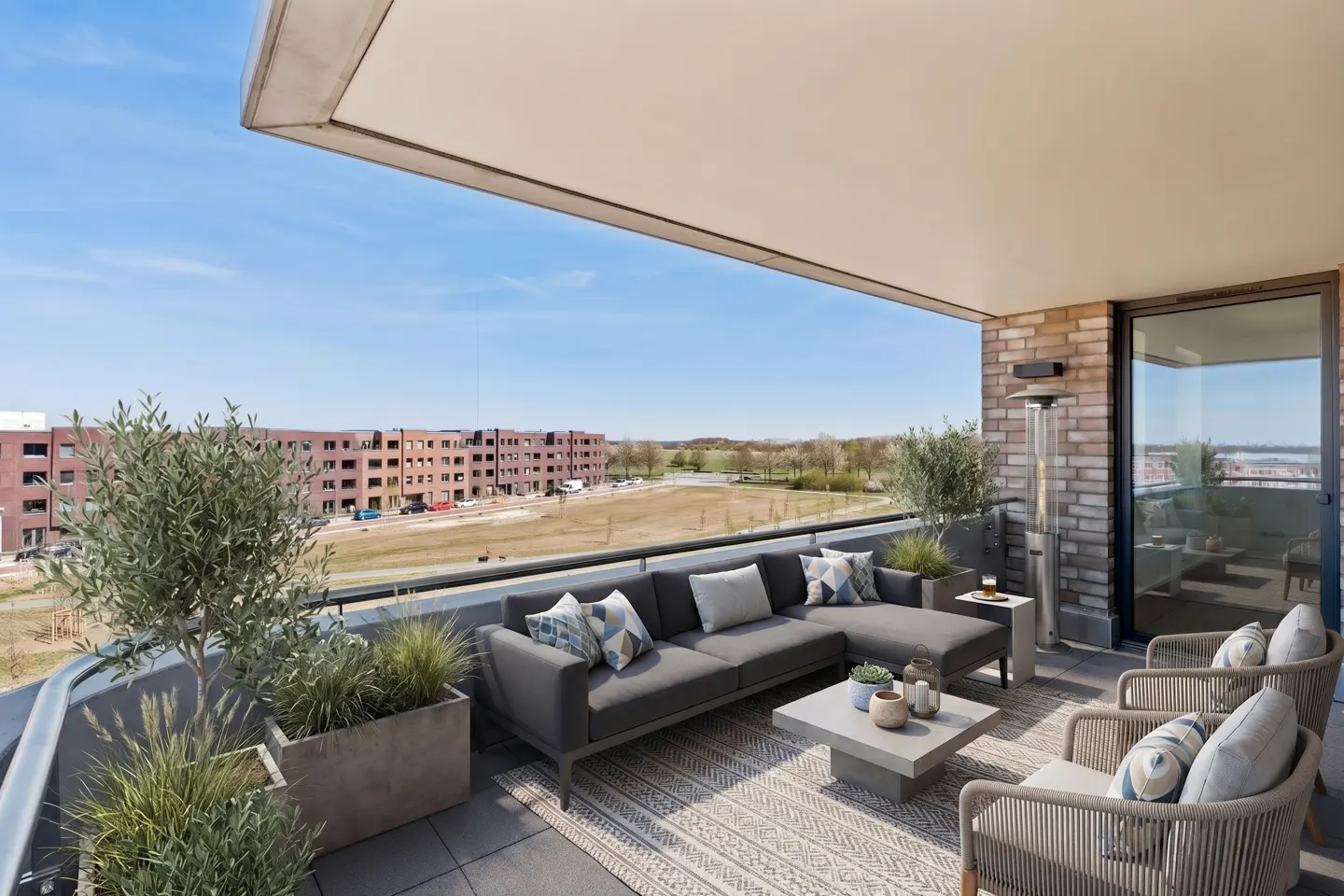 A modern balcony with gray sofa, chairs, and a table on a patterned rug. Potted plants add greenery, with a view of buildings and a blue sky in the background.