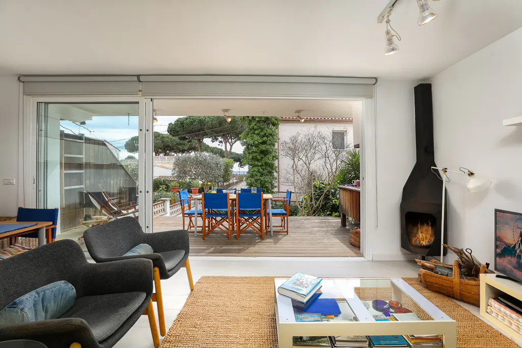 Modern living room with grey armchairs, glass coffee table, and fireplace, opening to a deck with blue dining chairs.