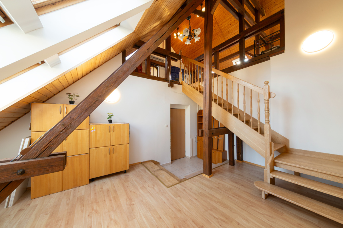 Attic room with wooden beams, light wood flooring, a staircase leading to a loft, and light wood storage cabinets. [AI-generated text]