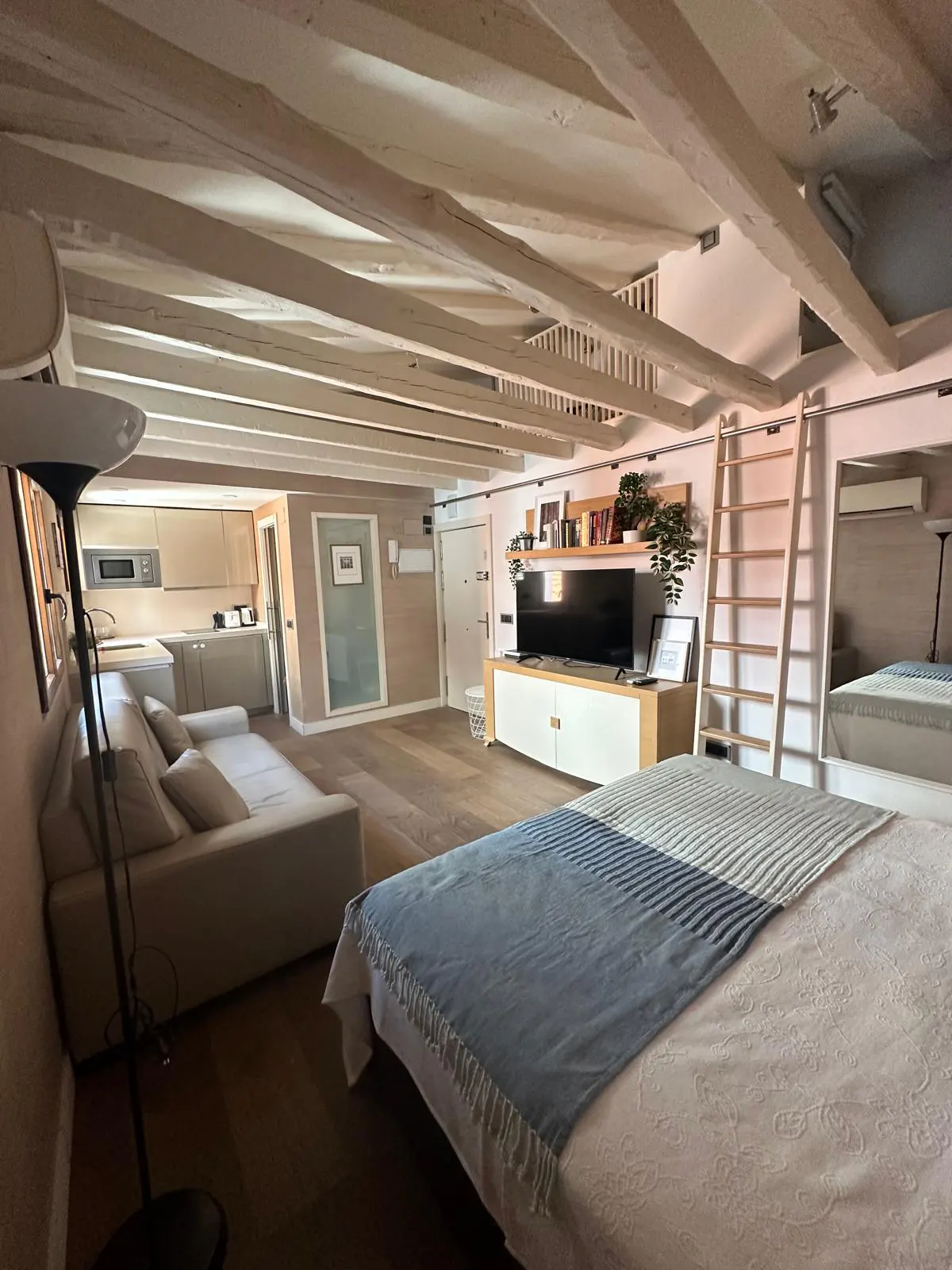 Modern studio apartment with exposed white ceiling beams, a bed with a blue throw, a sofa, and a kitchen in the background.