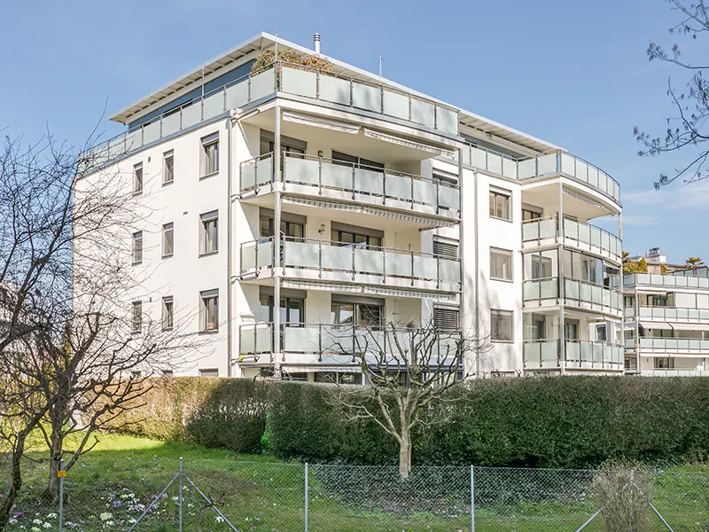 A modern, white apartment building with glass balconies and a rooftop terrace. Green lawn and bushes in front.
