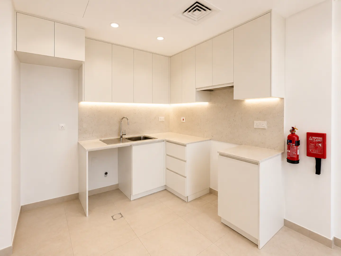 Modern kitchen with white cabinets, light stone countertops, under-cabinet lighting, a sink, and a wall-mounted fire alarm.