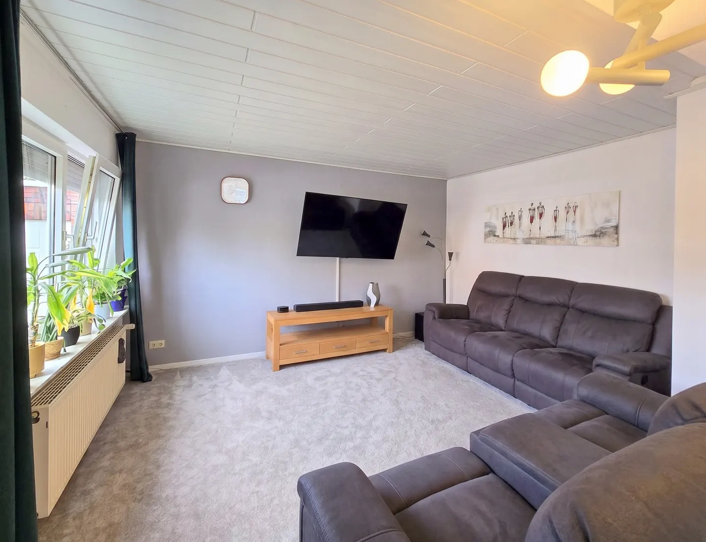 Living room with grey carpet, large dark grey sectional sofa, wall-mounted TV, wooden media console, and window plants.