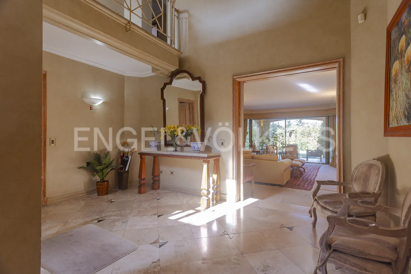 Foyer with marble floors, beige walls, and a wooden table with flowers. Two chairs face an open doorway to a living room with a view of greenery.