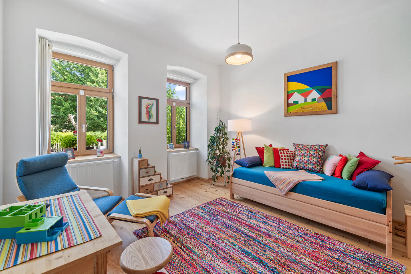 Bright living room with wood floors, white walls, and colorful accents. A blue daybed, chair, and a vibrant rug add pops of color.