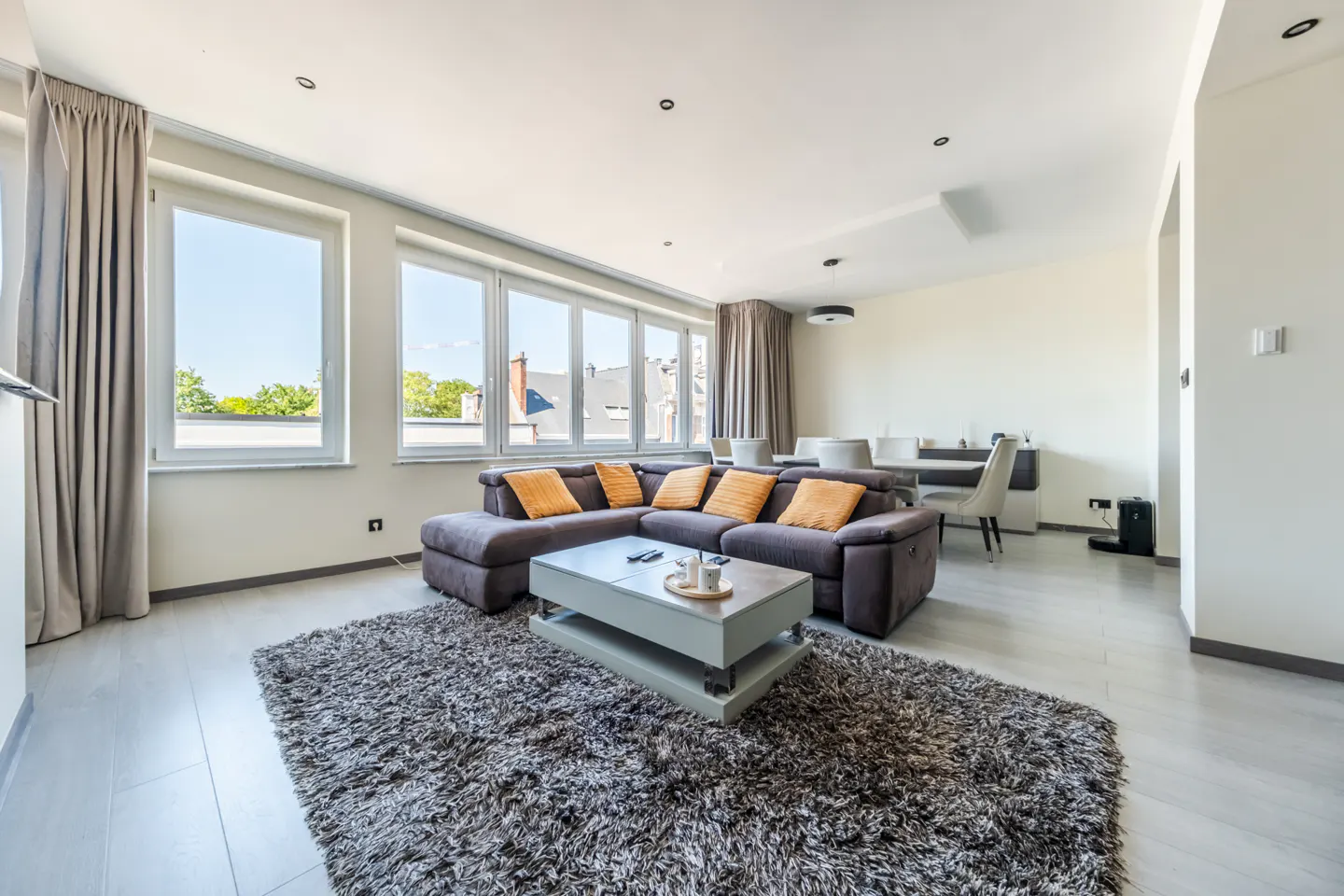 Bright living room with a gray sectional sofa, orange pillows, and a gray shag rug. Large windows offer a view of the outdoors.