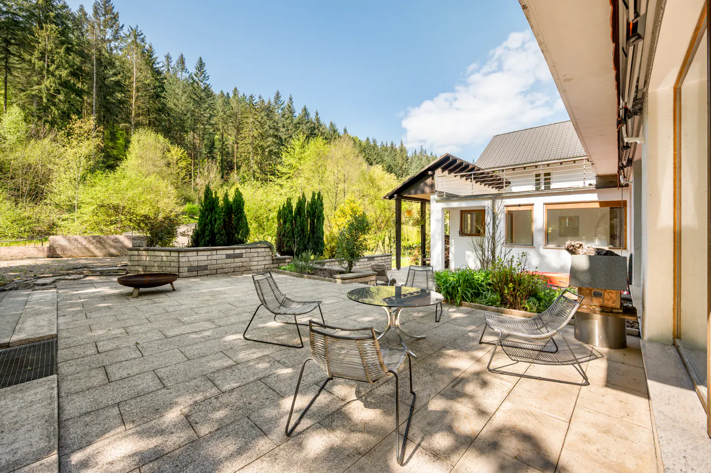 Outdoor patio with stone pavers, metal chairs, glass table, and forest backdrop on a sunny day.