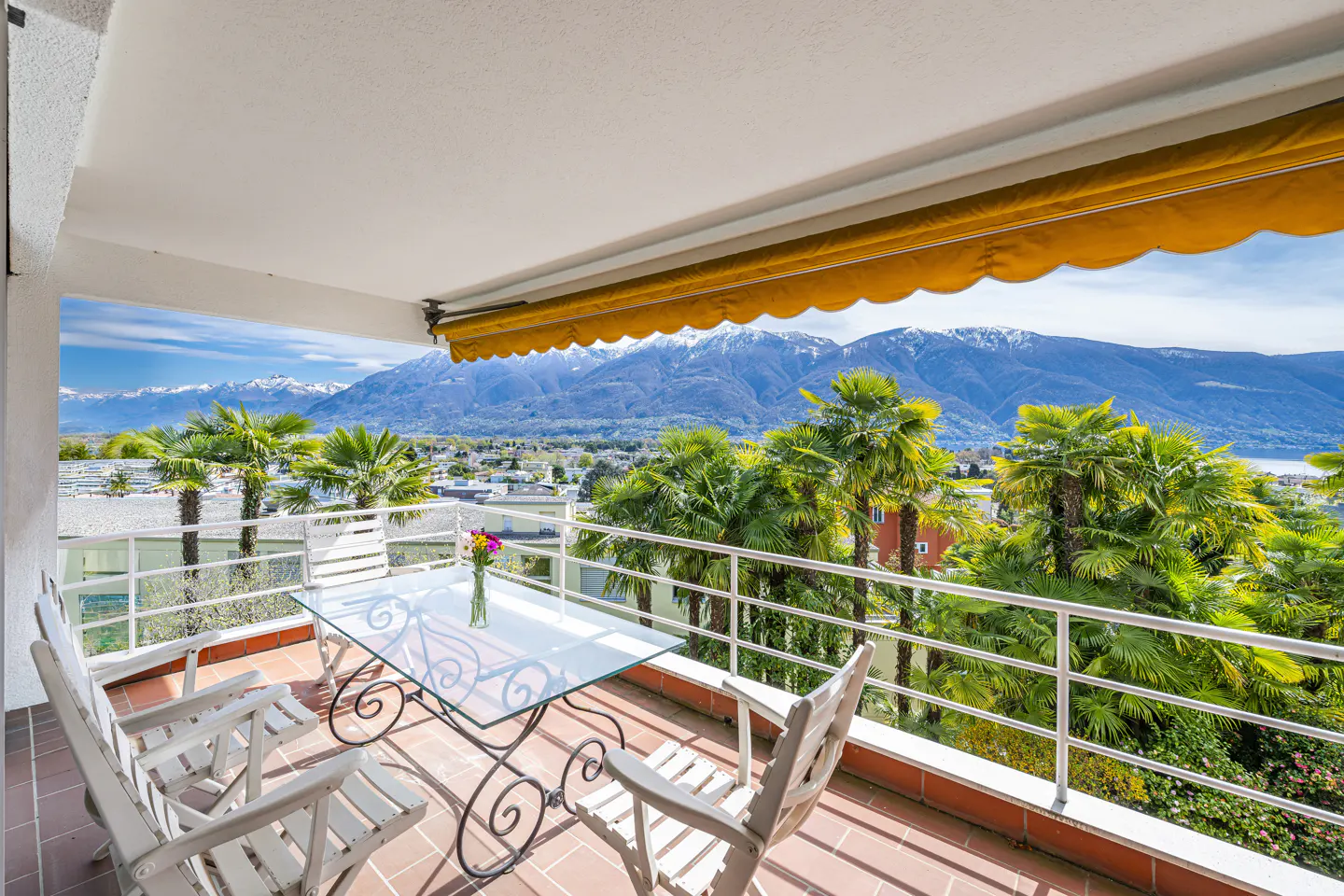 Balcony with a glass table, white chairs, and flowers. Palm trees and snow-capped mountains are in the background.