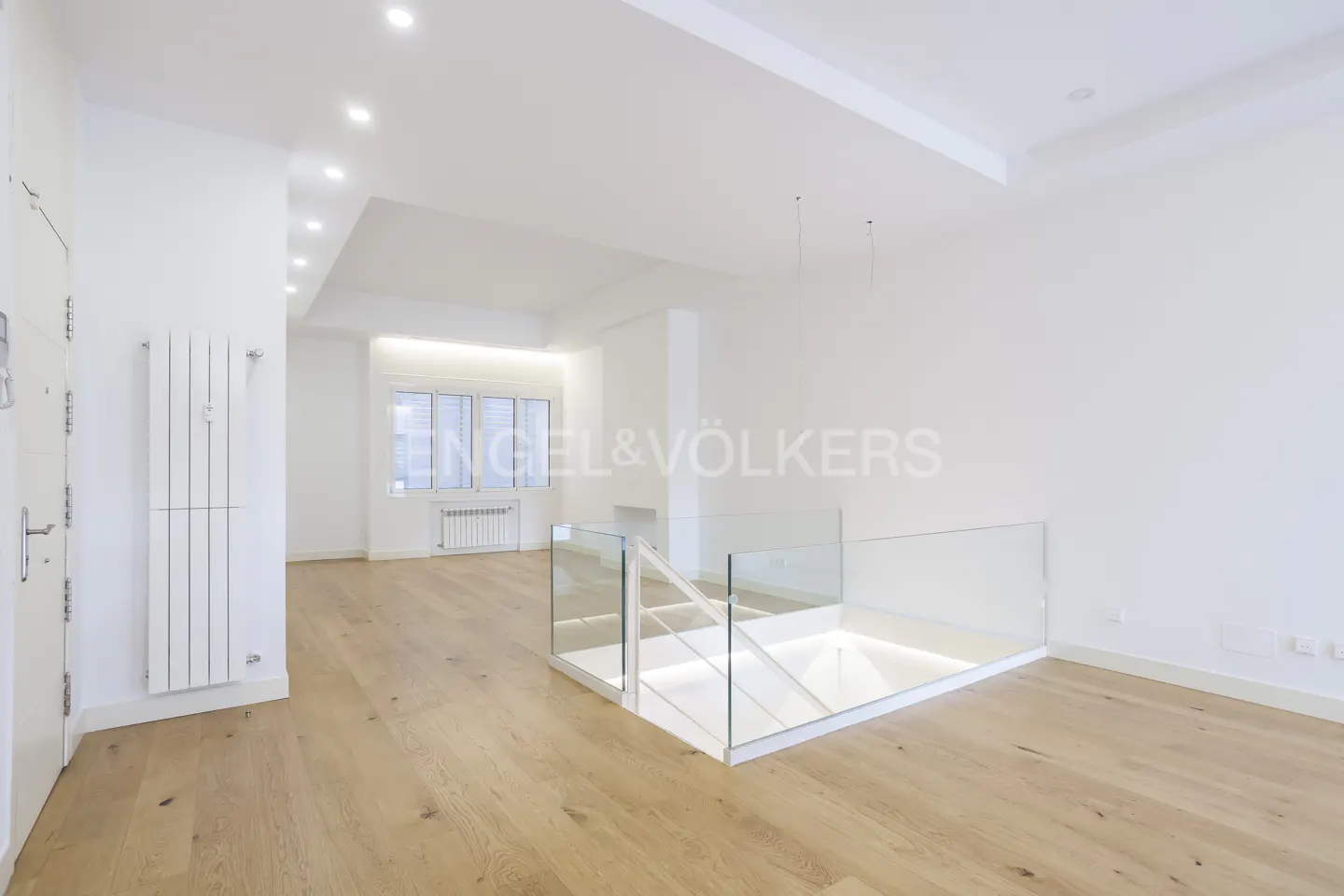 Bright, modern room with light wood floors, white walls, and recessed lighting. Glass-railed staircase leads down. Window in the background.