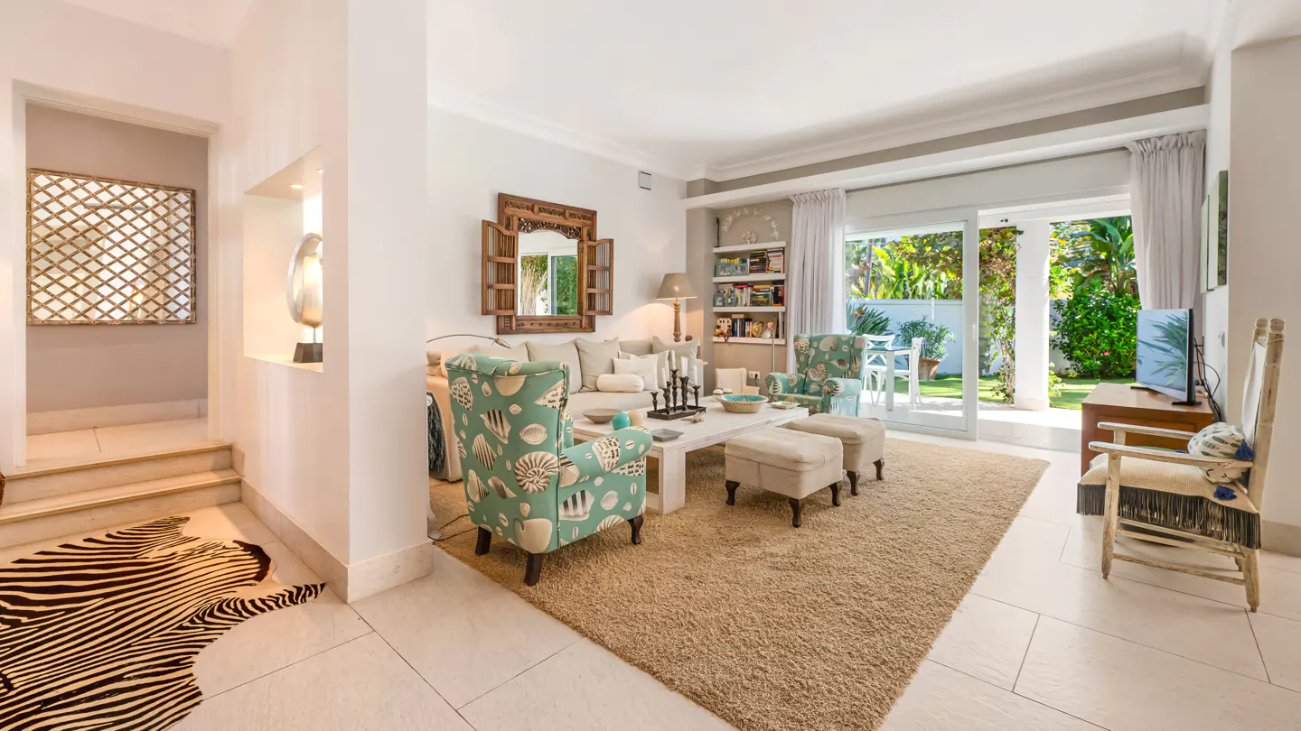 Bright living room with a beige rug, white sofa, and seashell-patterned chairs. Sliding glass doors lead to a green garden. A zebra-print rug is in the hall.