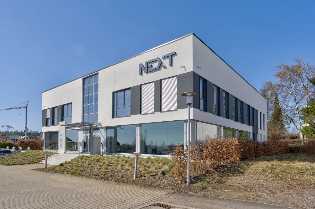 Exterior view of a modern, two-story office building with a "NEXT" sign, gray accents, and large windows.