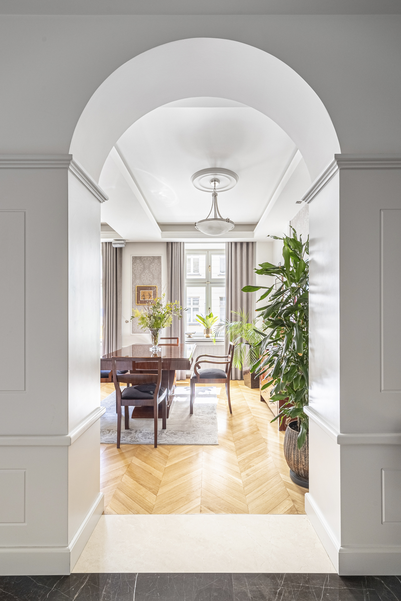 View through an arched doorway into a dining room with a wood table, chairs, and herringbone wood floors. A large potted plant sits to the right.