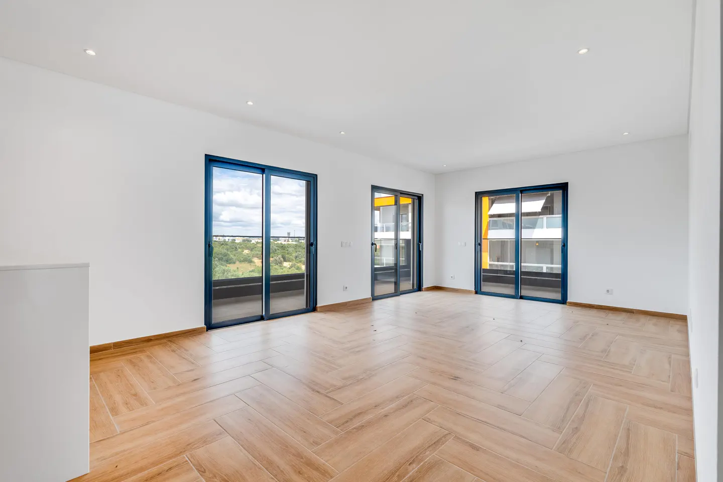 Bright, empty room with white walls, herringbone wood floors, and three blue-framed sliding glass doors to balconies.