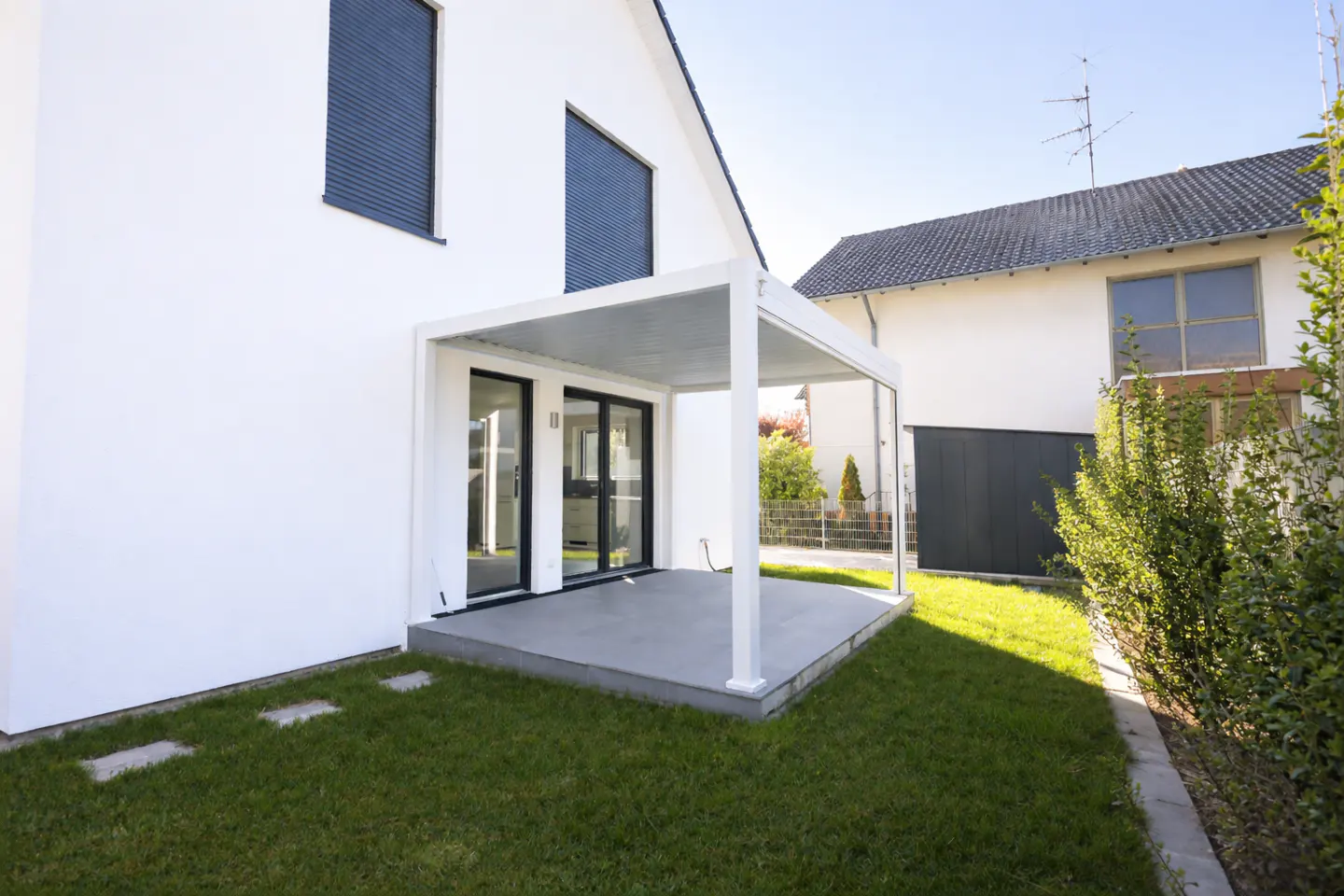 Exterior of a modern white house with a white pergola and black framed sliding glass doors. Green lawn and blue window shades.