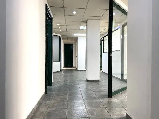 Hallway with gray tile floor, white walls, and dark green door frames. Recessed lighting in the ceiling. Glass-walled offices on the right.