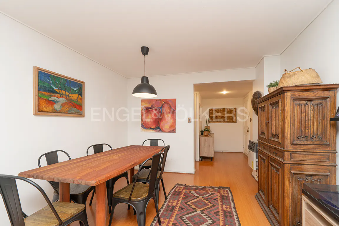 A dining room with a wooden table, black chairs, and hardwood floors. Artwork adorns the walls, and a vintage cabinet stands in the background.