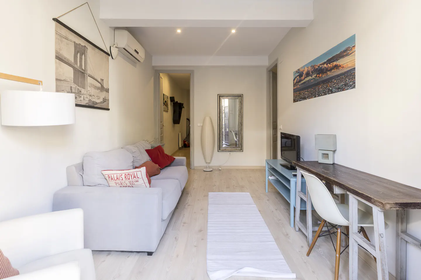 Small living room with a light gray sofa, wooden desk, white chair, and wall art in a bright, modern apartment.