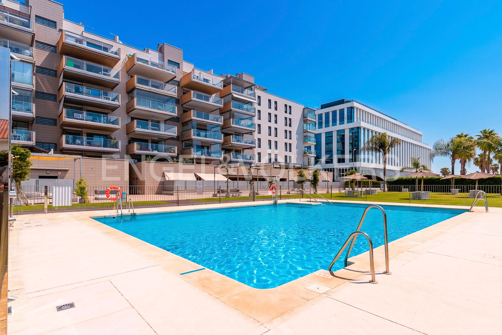 A bright blue swimming pool in front of a modern apartment complex with balconies under a clear sky.