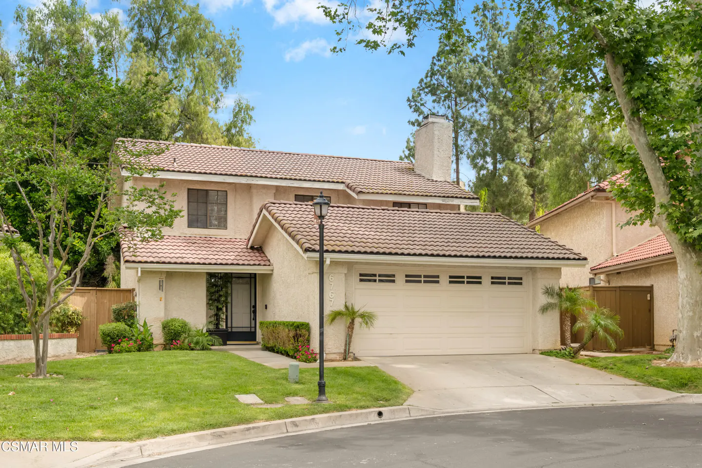 Two-story beige house with a brown tile roof, a two-car garage, and green lawn. Trees and blue sky in the background.