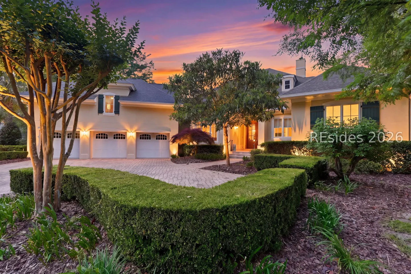Beige two-story house with a three-car garage, green shutters, and manicured hedges at sunset.