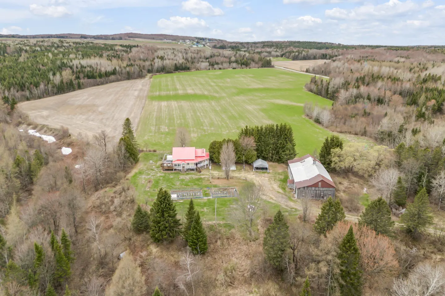 Aerial view of a rural property with a red-roofed house, barn, fields, and surrounding forest.