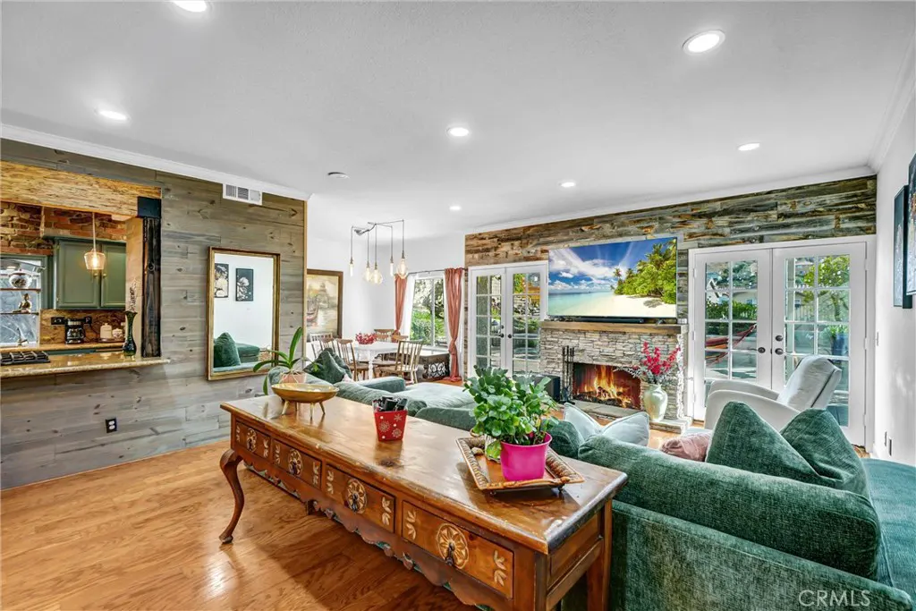 A living room with a green sofa, a wooden table, and a stone fireplace with a TV above it. The TV shows a beach scene. There is a dining table in the background.