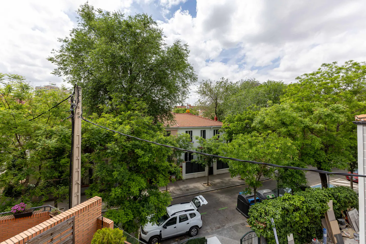 View from a brick balcony overlooking a street with trees, a white car with an open trunk, and a white house with a red tile roof.
