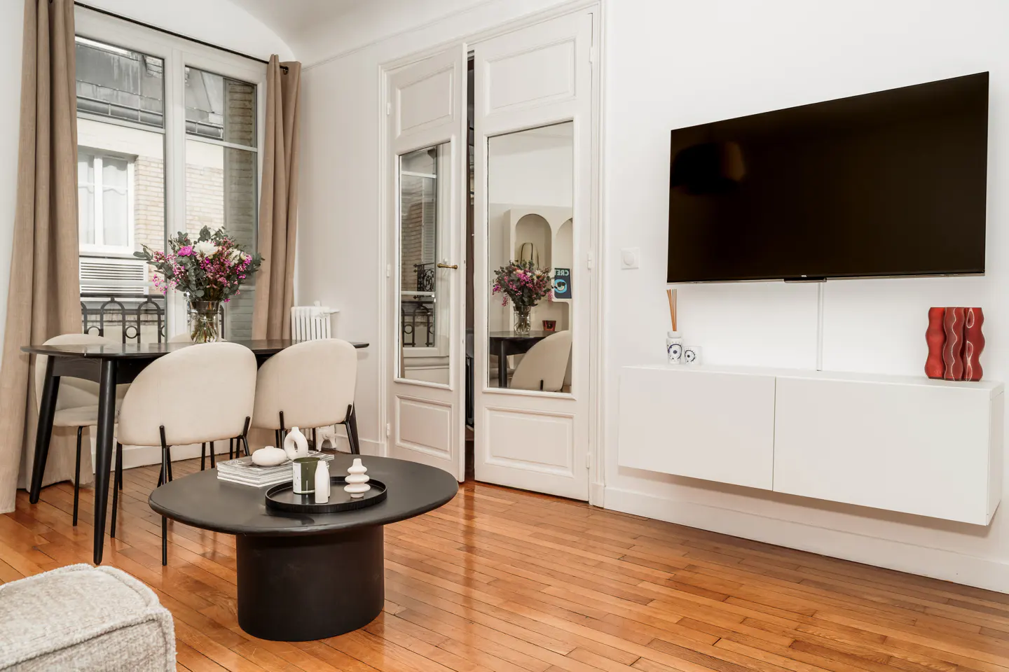 Bright living room with wood floors, white walls, and a large TV. A black table and chairs sit near a window with beige curtains. French doors reflect the room.