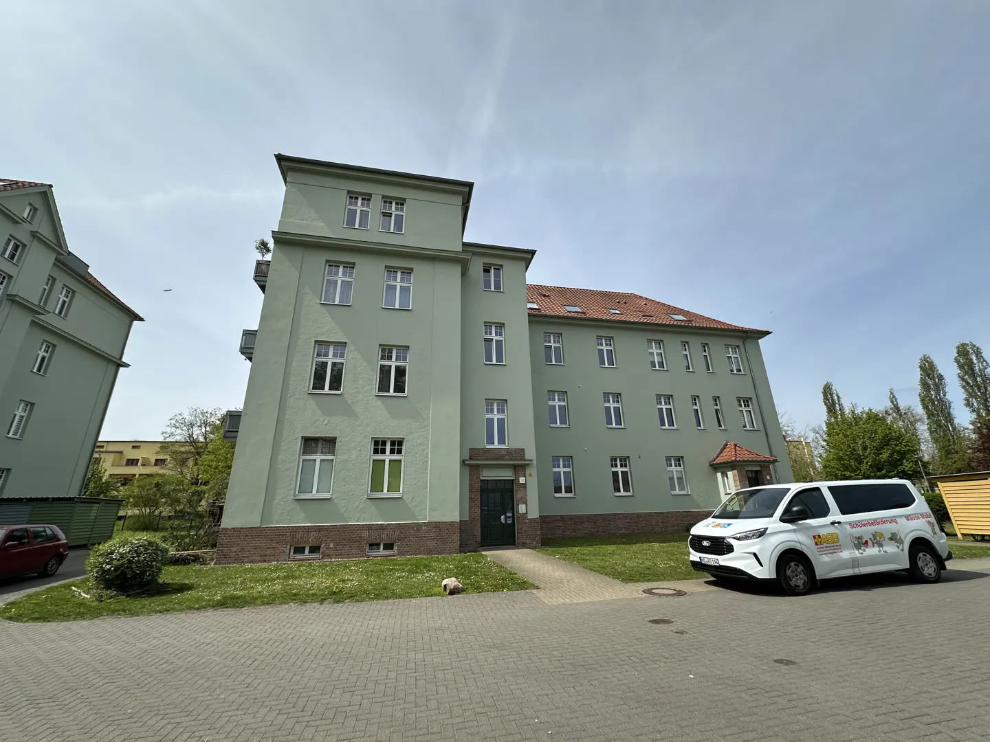A three-story, light green apartment building with white windows and a white van parked in front.