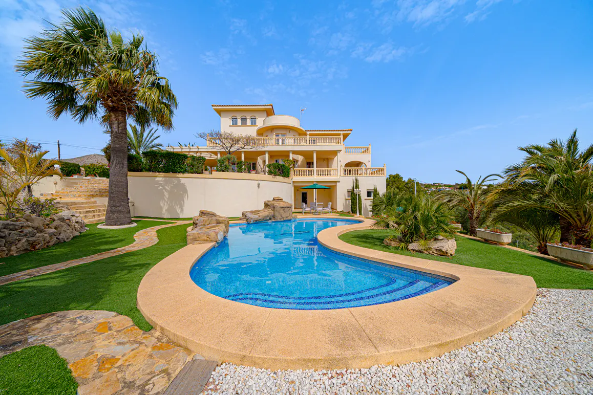 Exterior view of a multi-story beige house with a blue tiled pool, palm trees, and green lawn under a blue sky.