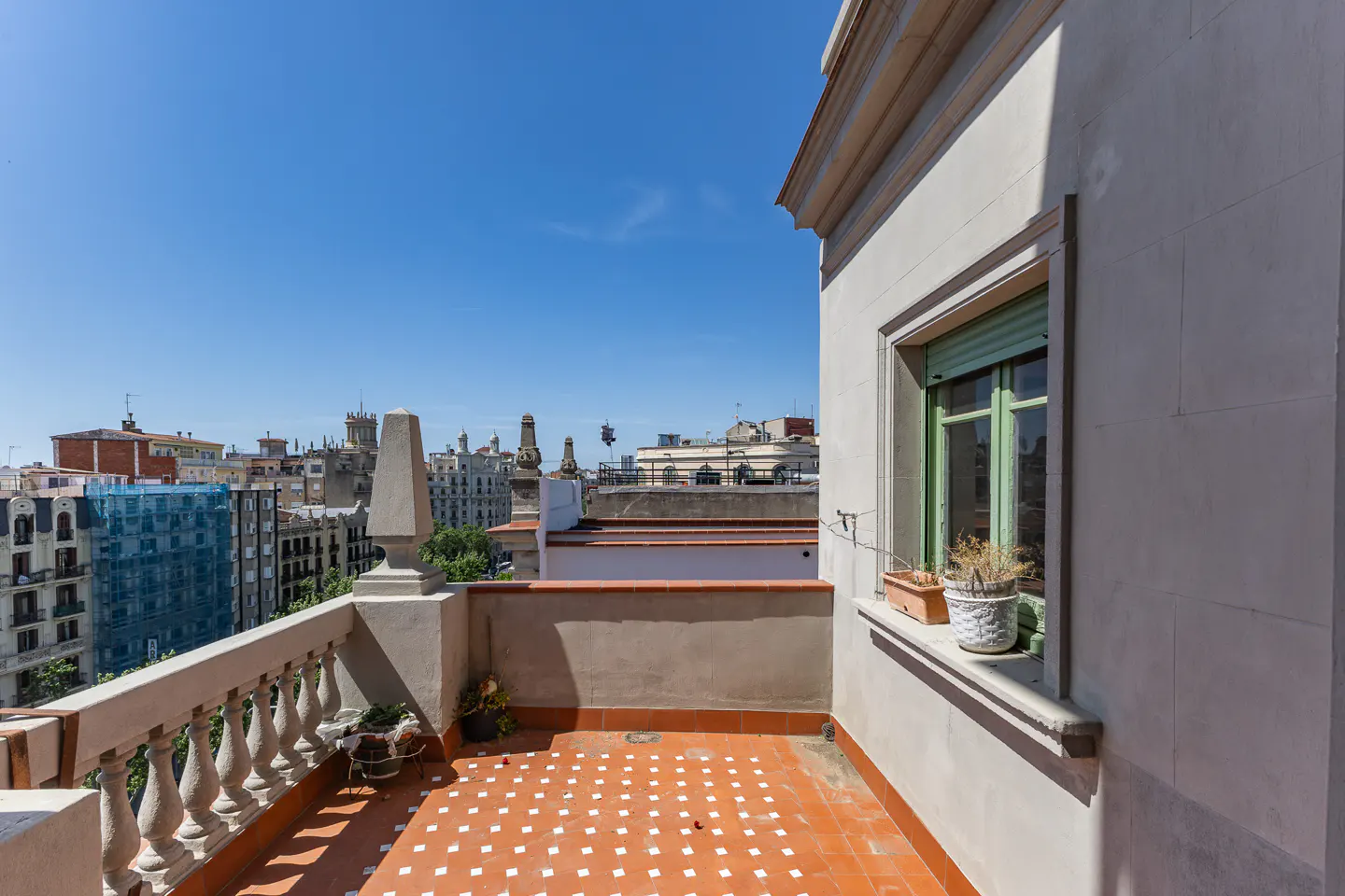 Balcony with red tile floor and white railing overlooks a city skyline under a clear blue sky. A window with green trim is on the right.