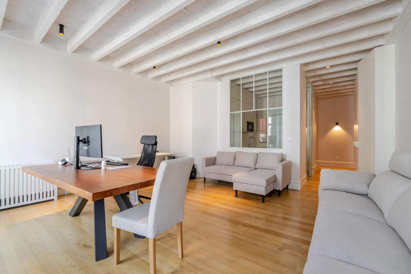 Bright, modern living space with wood floors, white walls, and beamed ceiling. A desk, sofas, and a glimpse of a kitchen are visible.