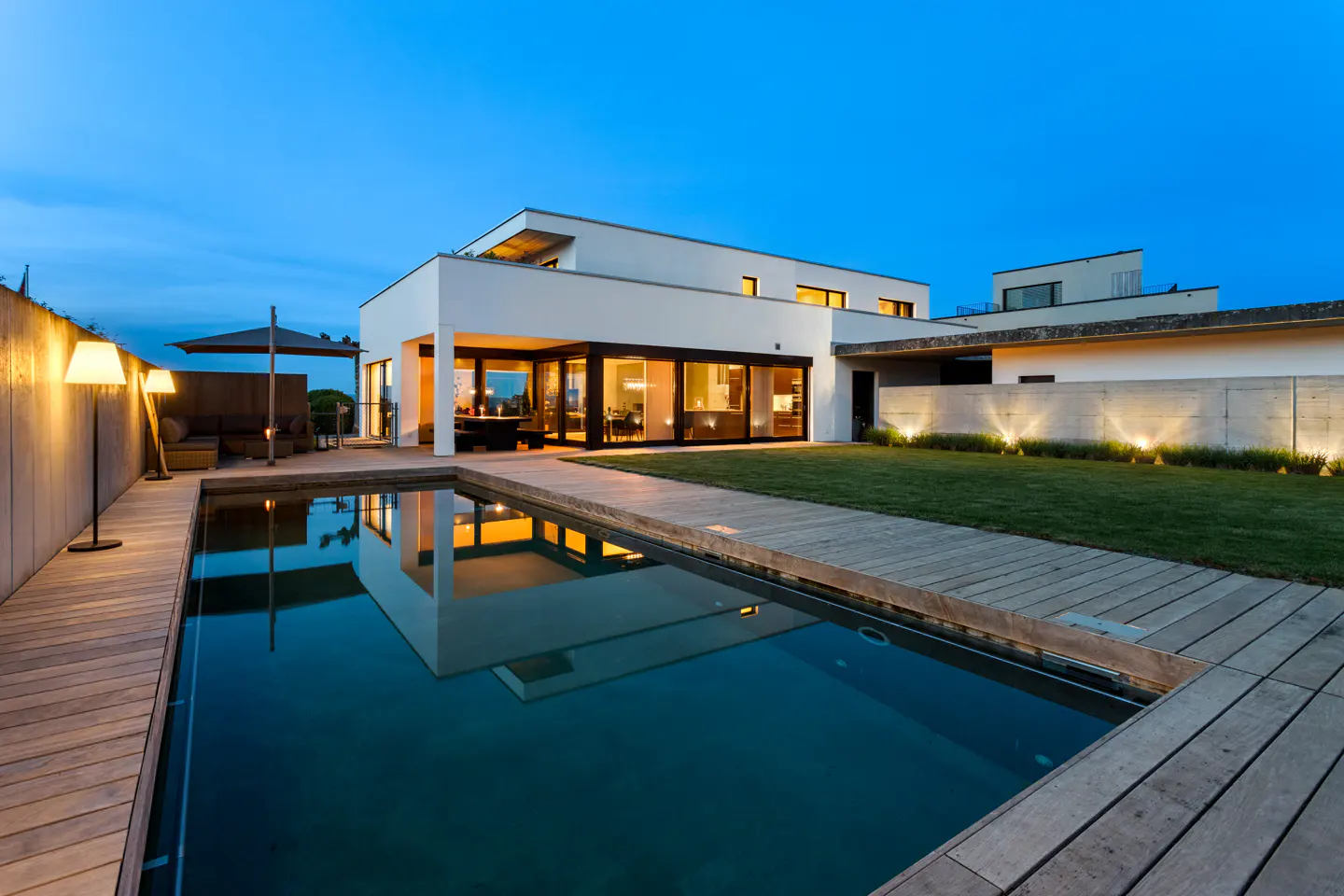 Modern white house with a pool, wooden deck, and green lawn at dusk. Lights illuminate the yard and reflect in the water.