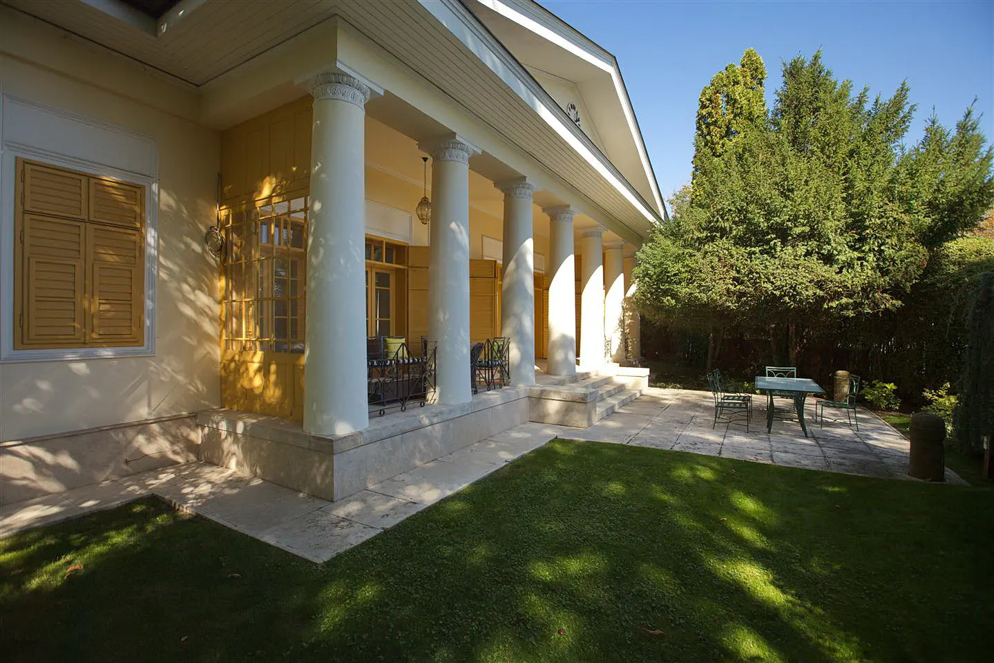 Exterior view of a yellow house with white columns and a green lawn. A patio set sits on a stone patio.