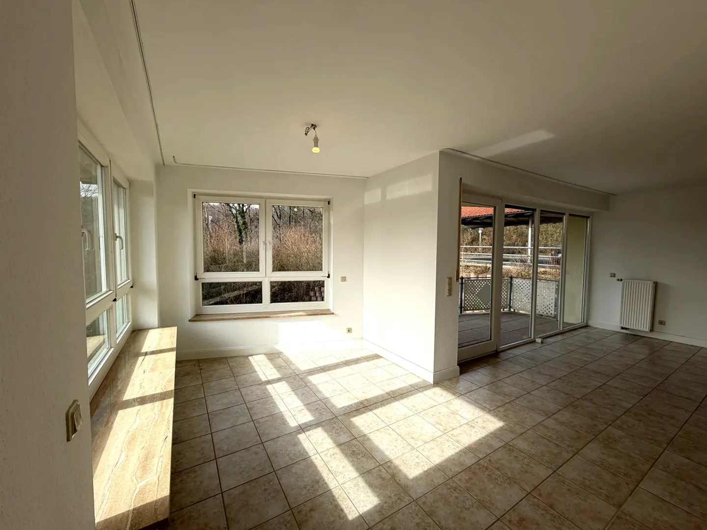 Bright, empty room with tile floor, white walls, and large windows. Sunlight streams in, creating shadows. Sliding glass doors lead to a balcony.