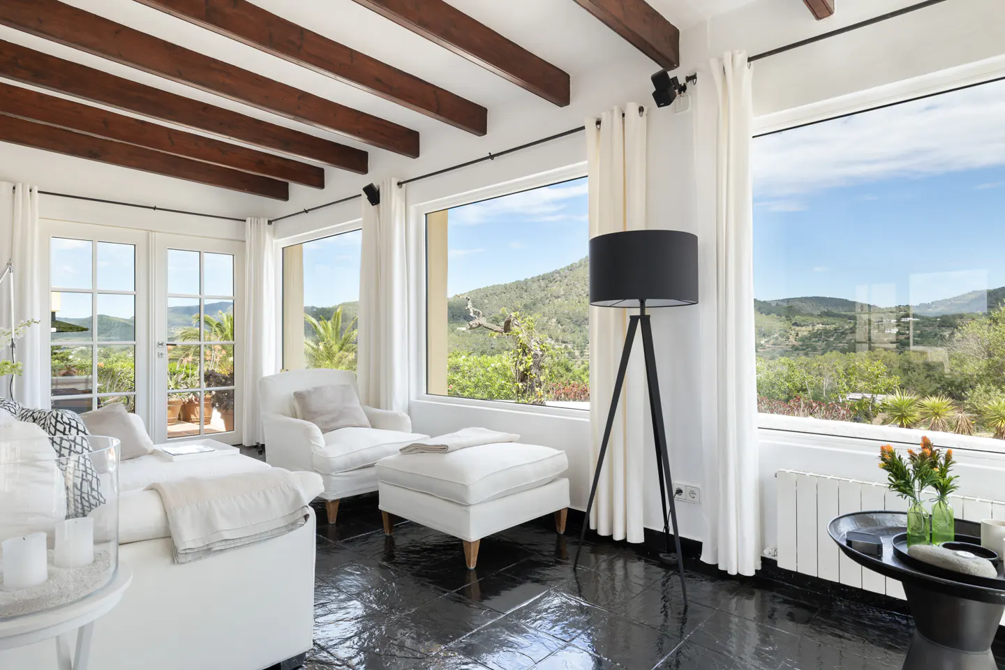 Bright living room with white furniture, black floors, and exposed wood beams. Large windows offer a view of lush greenery.