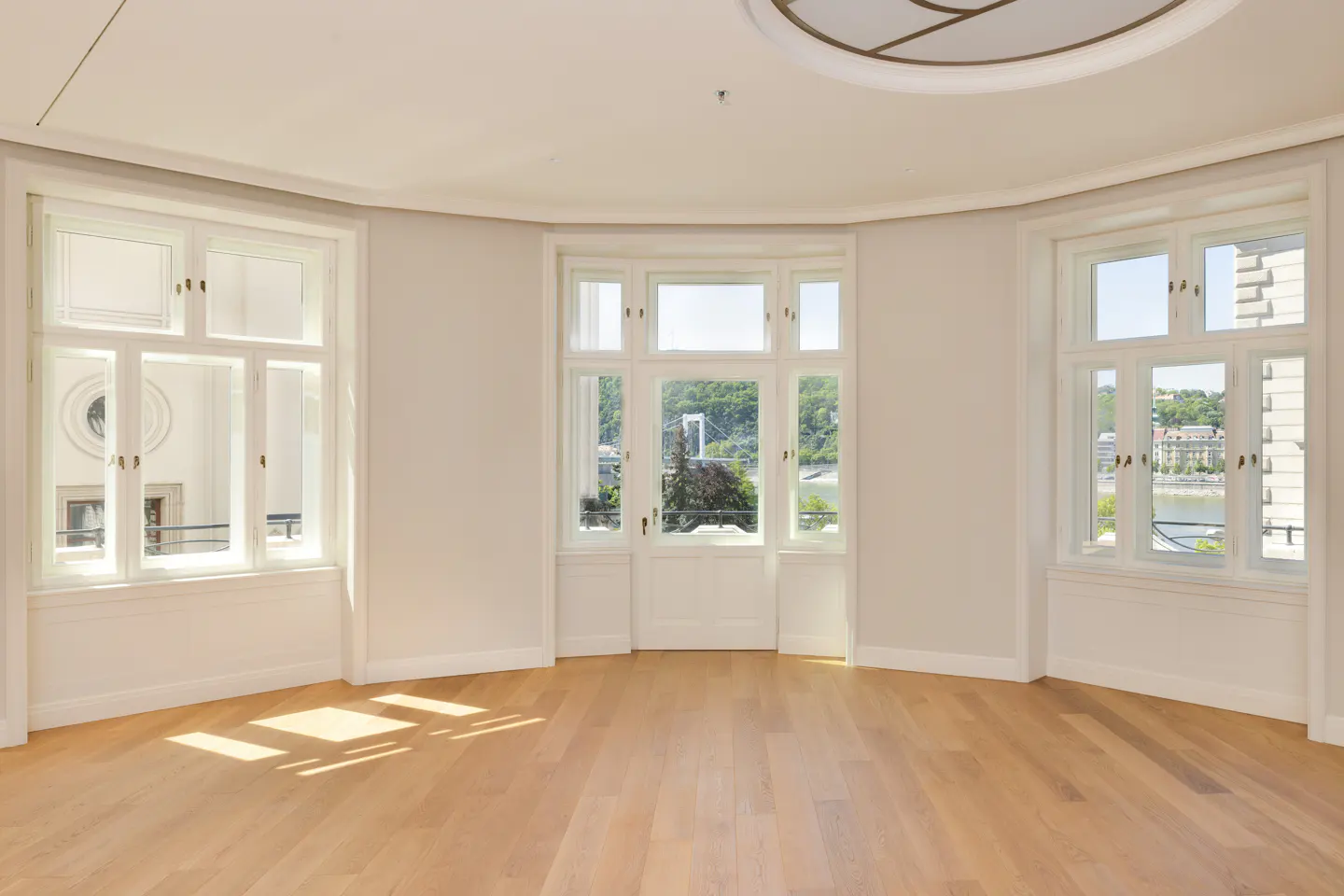 Bright, empty room with light wood floors, white walls, and three large windows. The center window has a view of a bridge and greenery.