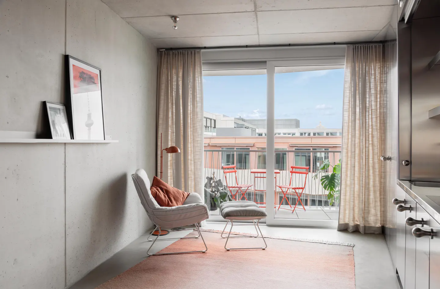 Modern apartment interior with concrete walls, a chair, and a balcony with red chairs and a city view.