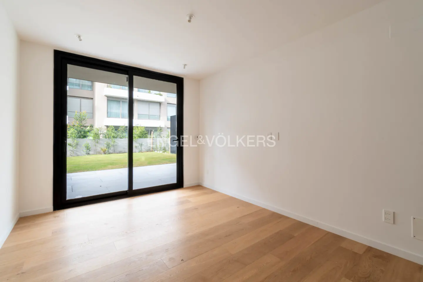 Empty room with light wood floors, white walls, and a large sliding glass door opening to a private patio and garden.