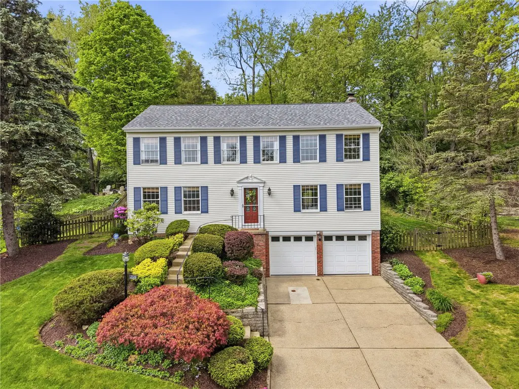 Two-story house with white siding, blue shutters, and a red door. Landscaped front yard with colorful bushes and a two-car garage.