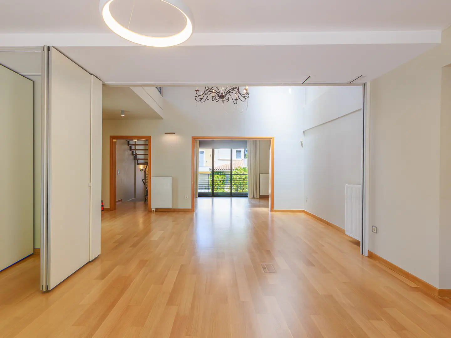 Bright, empty room with light wood floors, white walls, and a modern chandelier. A staircase and balcony are visible in the background.