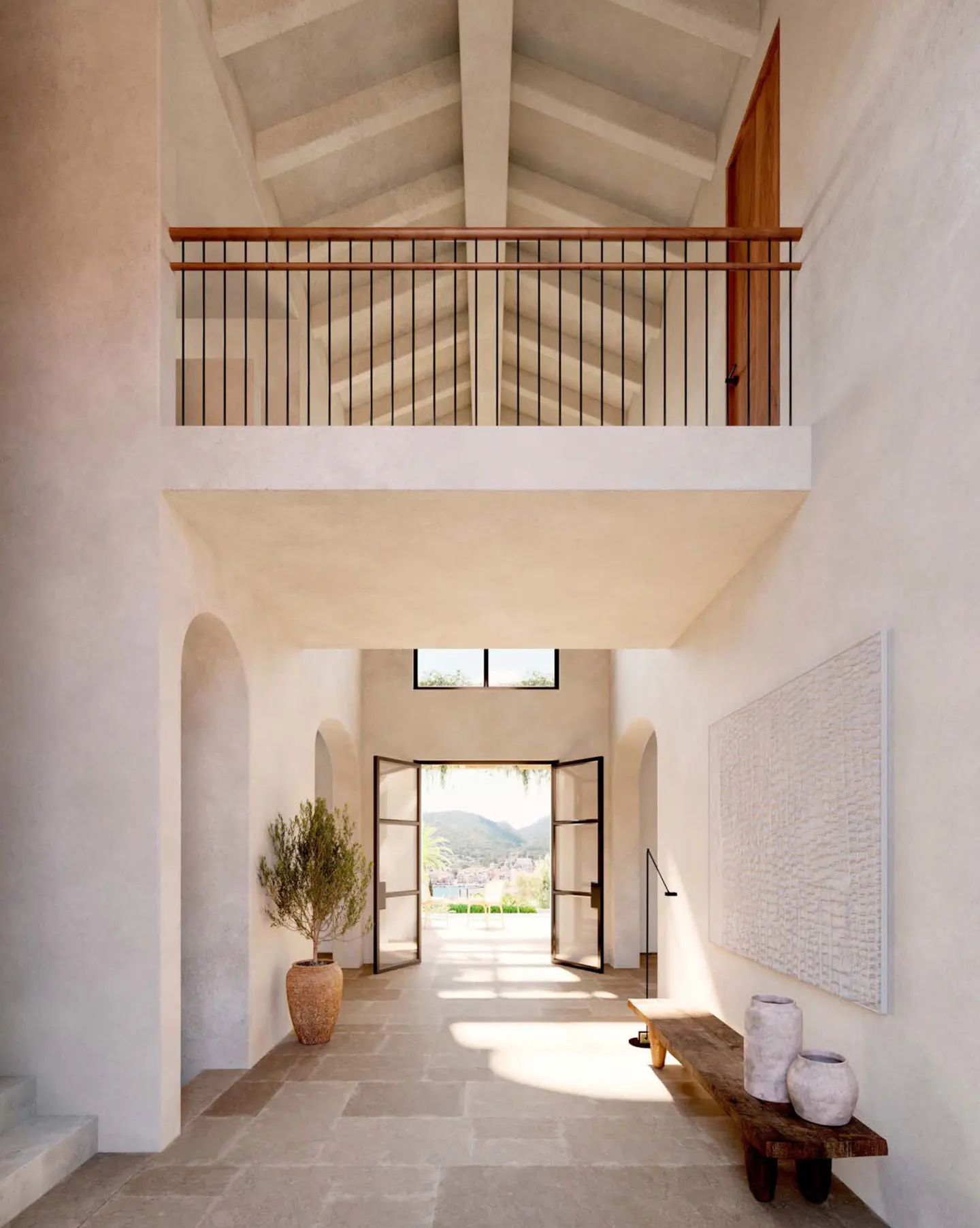 Interior view of a bright foyer with stone floors, white walls, and a balcony overlooking the space. French doors open to a view of hills.