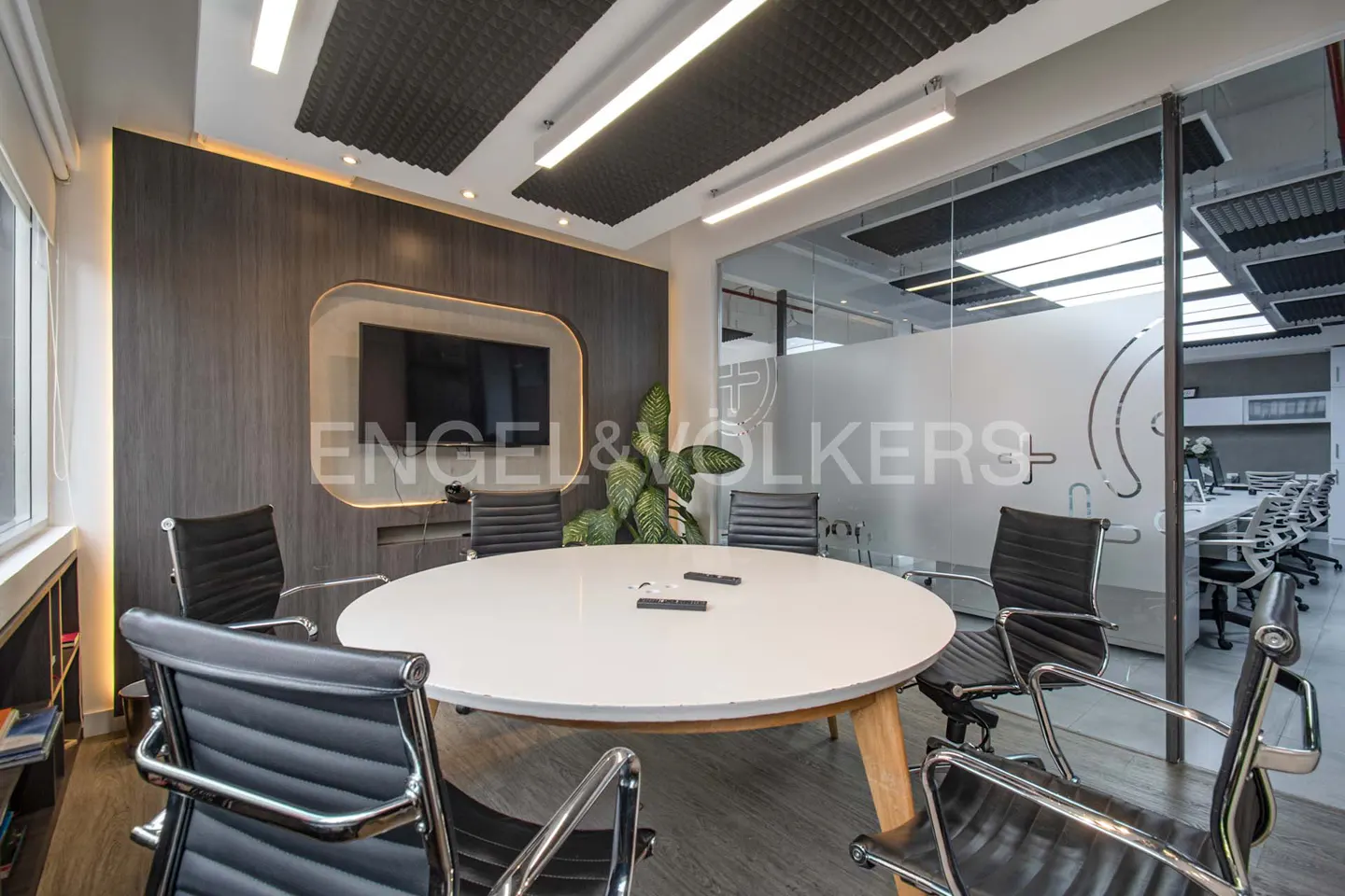 A modern conference room with a white round table, black chairs, and a large screen on a wood-paneled wall.