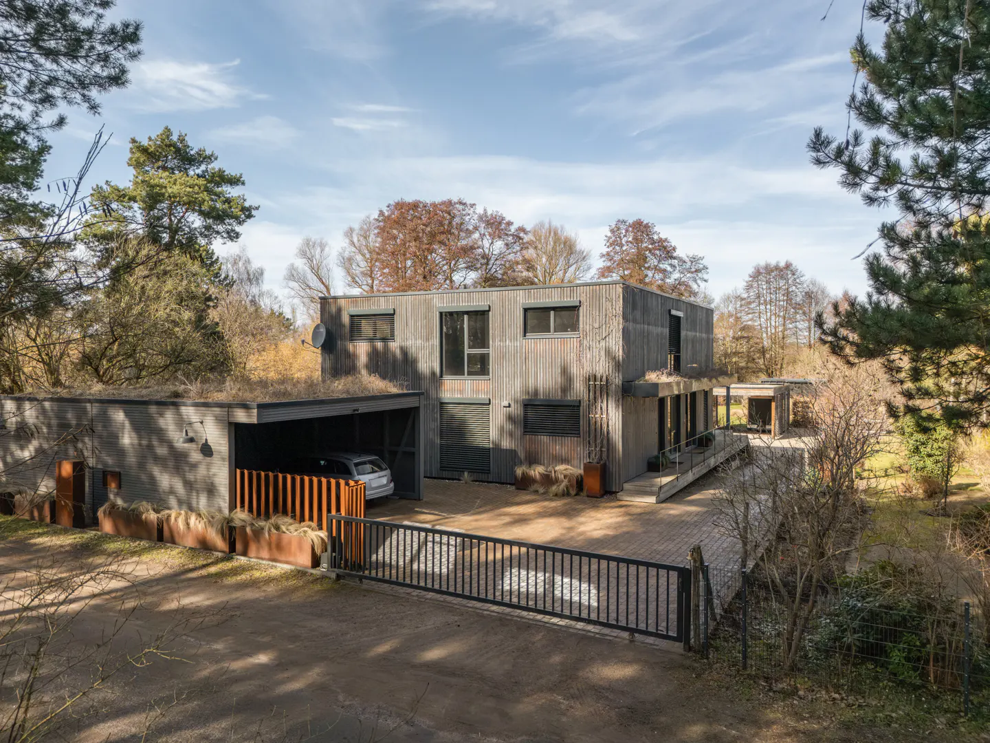 Two-story modern home with gray wood siding, a green roof on the garage, and a black metal gate. A car is parked in the garage.