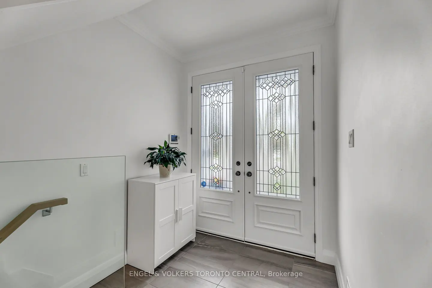 Bright foyer with white walls, double doors with decorative glass, and a white cabinet with a potted plant.