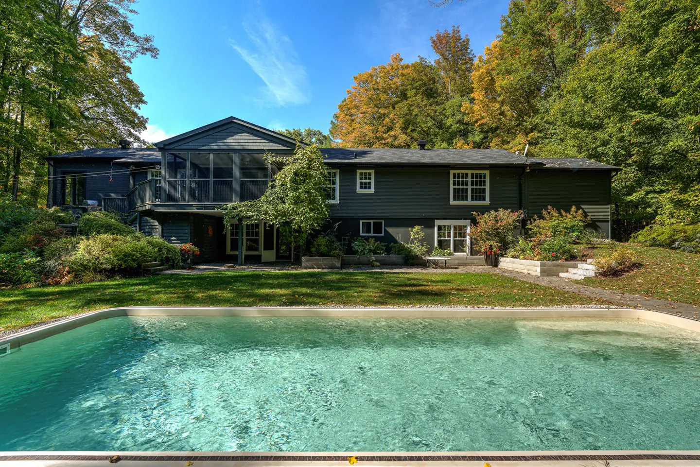 Backyard view of a dark gray house with a pool, green lawn, and trees under a blue sky.